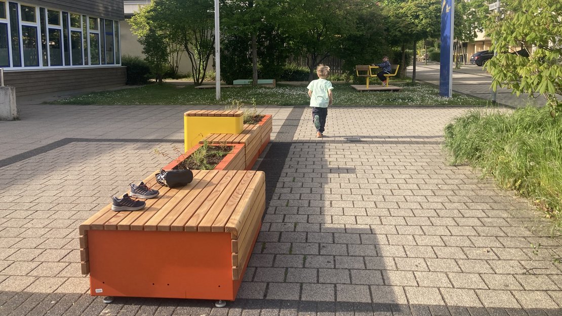 Eine mit Holzleisten verkleidete orange- und gelbfarbene Sitz-/Pflanzkombination steht auf einem gepflasterten Untergrund. Links im Bild befindet sich die Fensterfront eines Gebäudes. Rechts ist ein verkehrsberuhigter Bereich mit einem parkenden Auto zu sehen. Im Hintergrund, auf einer Rasenfläche im Schatten, steht eine gelbe Sitzgruppe. Zwei Kinder spielen bei sonnigem Wetter.